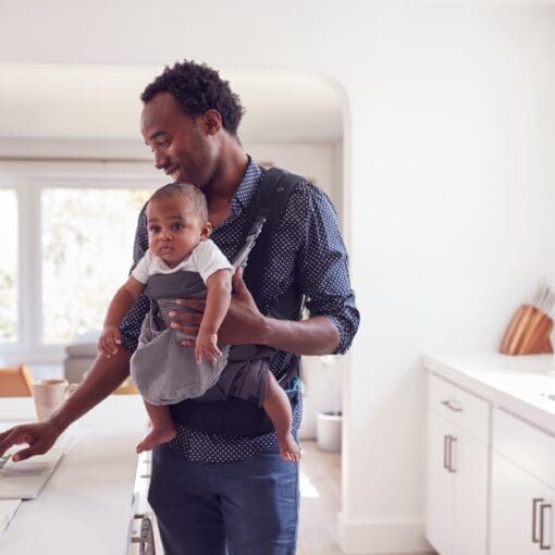 Father With Baby Daughter In Sling Multi-Tasking Working From Home On Laptop