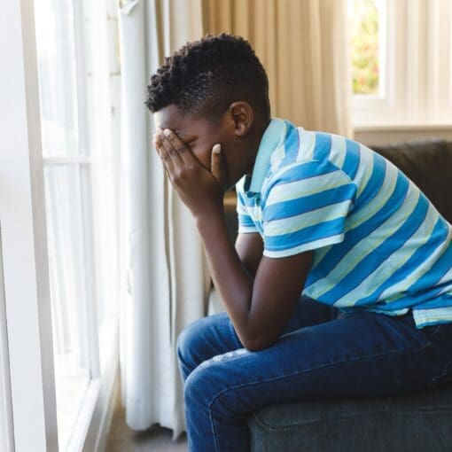 Sad African American Boy Covering His Face And Sitting At Window In Living Room