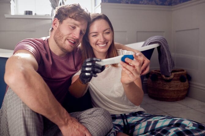 Excited Couple With Woman With Prosthetic Arm Sitting On Bathroom Floor With Positive Pregnancy Test